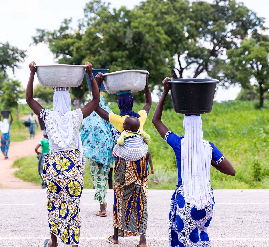 Women wearing colorful fress walking away from the camera holding buckets of water on their heads